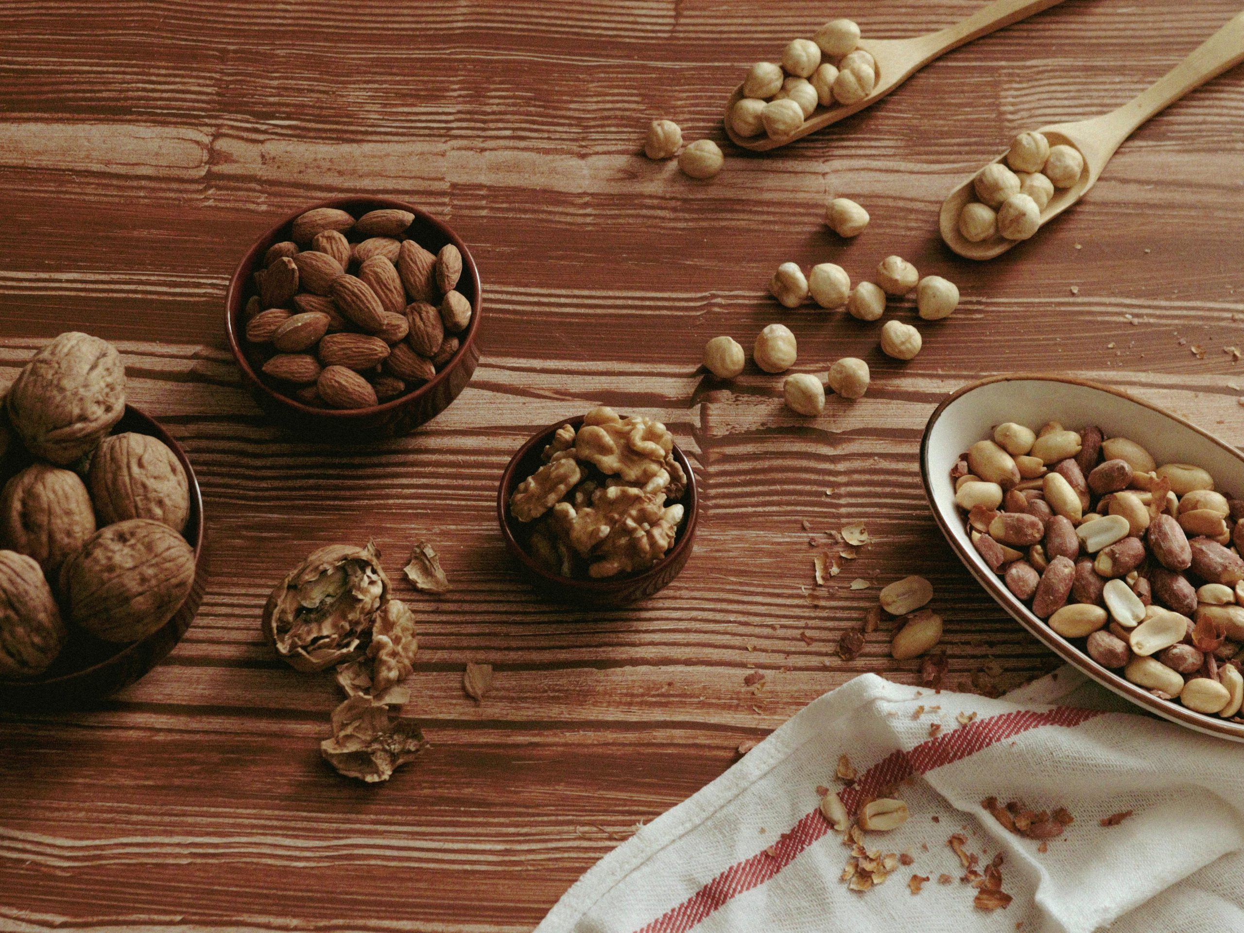 Our Products A top view of various nuts in bowls and spoons on a wooden tabletop.